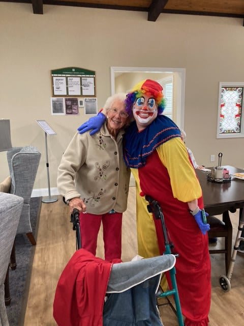 An elderly person and a clown share a friendly embrace indoors.