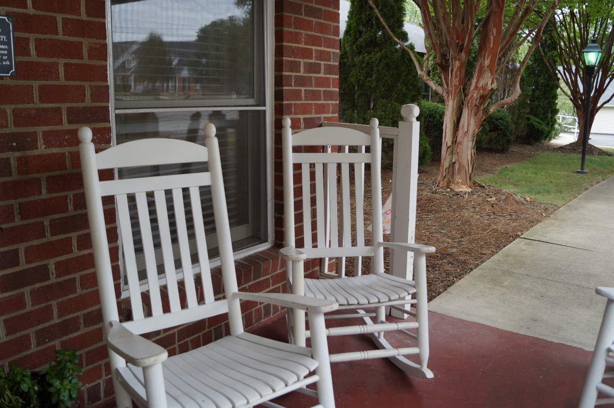 Two white rocking chairs on a porch outside a brick house.