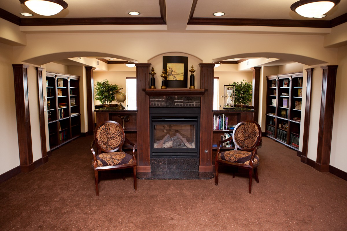 A cozy library interior with a fireplace, flanked by two chairs and surrounded by bookshelves.