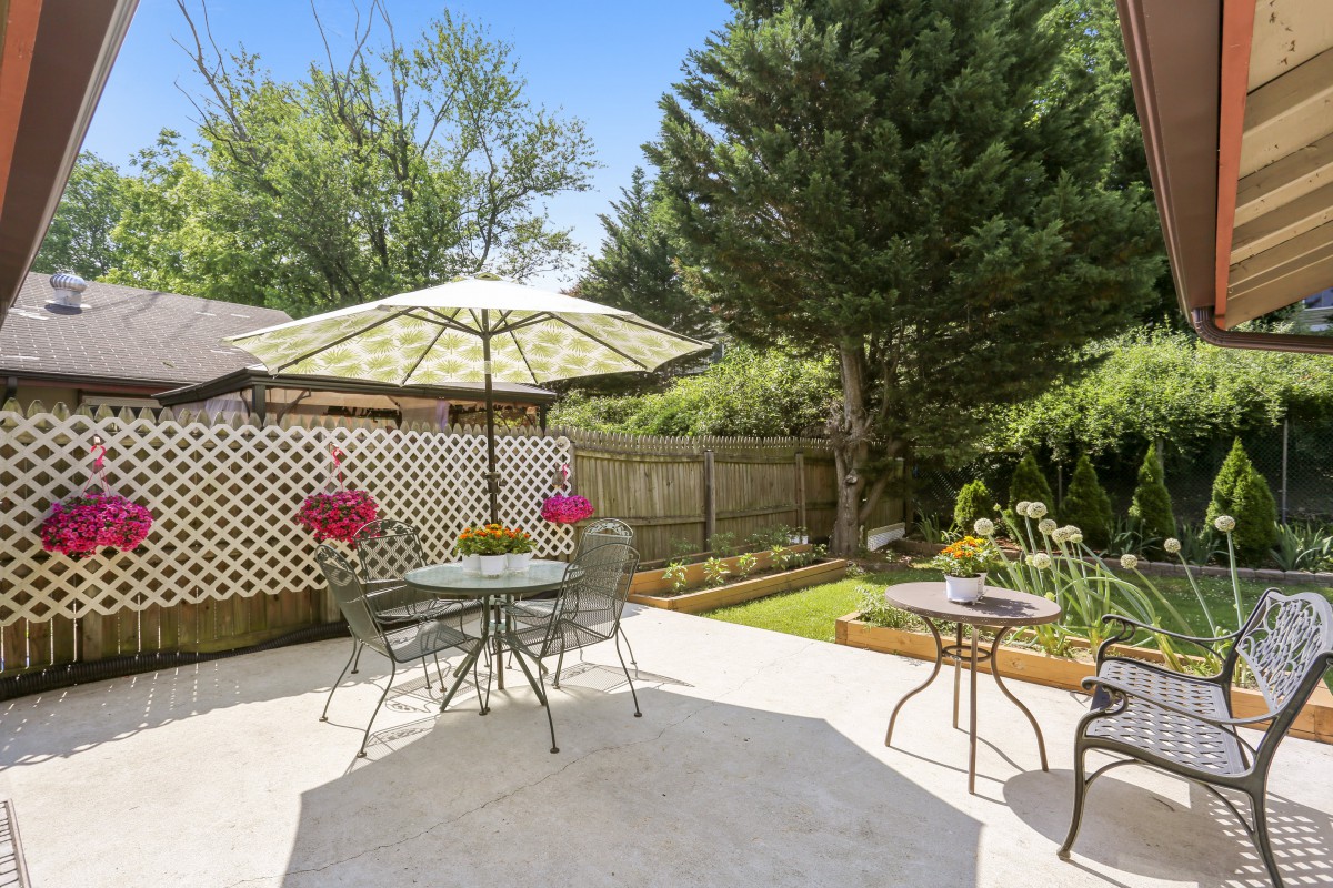 A sunny backyard patio featuring outdoor furniture with an umbrella, surrounded by a lush garden and fence.