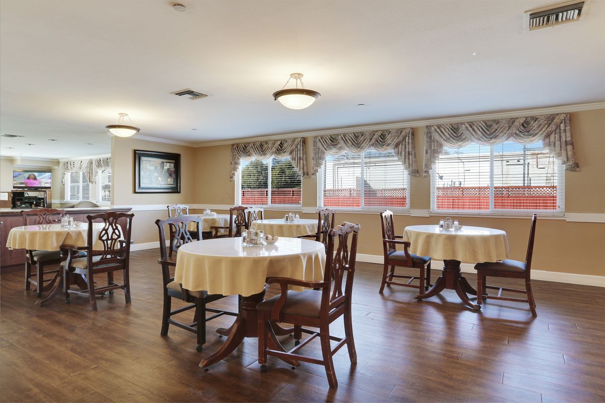 A cozy dining area with neatly arranged tables and chairs, wooden flooring, and large windows dressed with curtains.