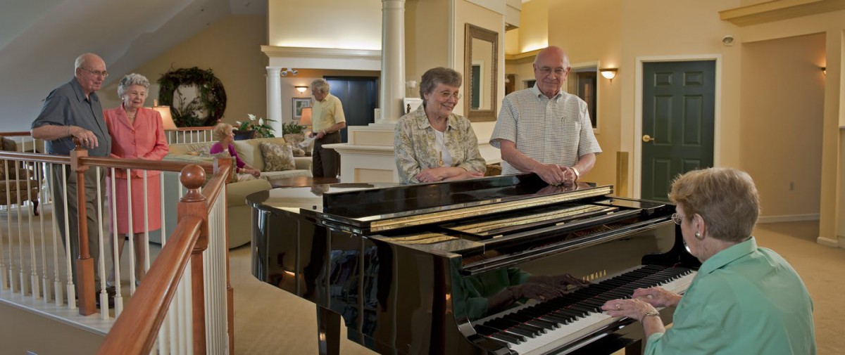 An elderly couple standing beside a grand piano in a well-furnished living room.