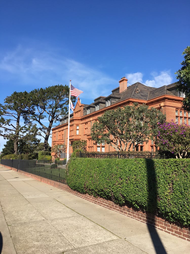 A large brick mansion with a manicured hedge and an American flag flying on a sunny day.