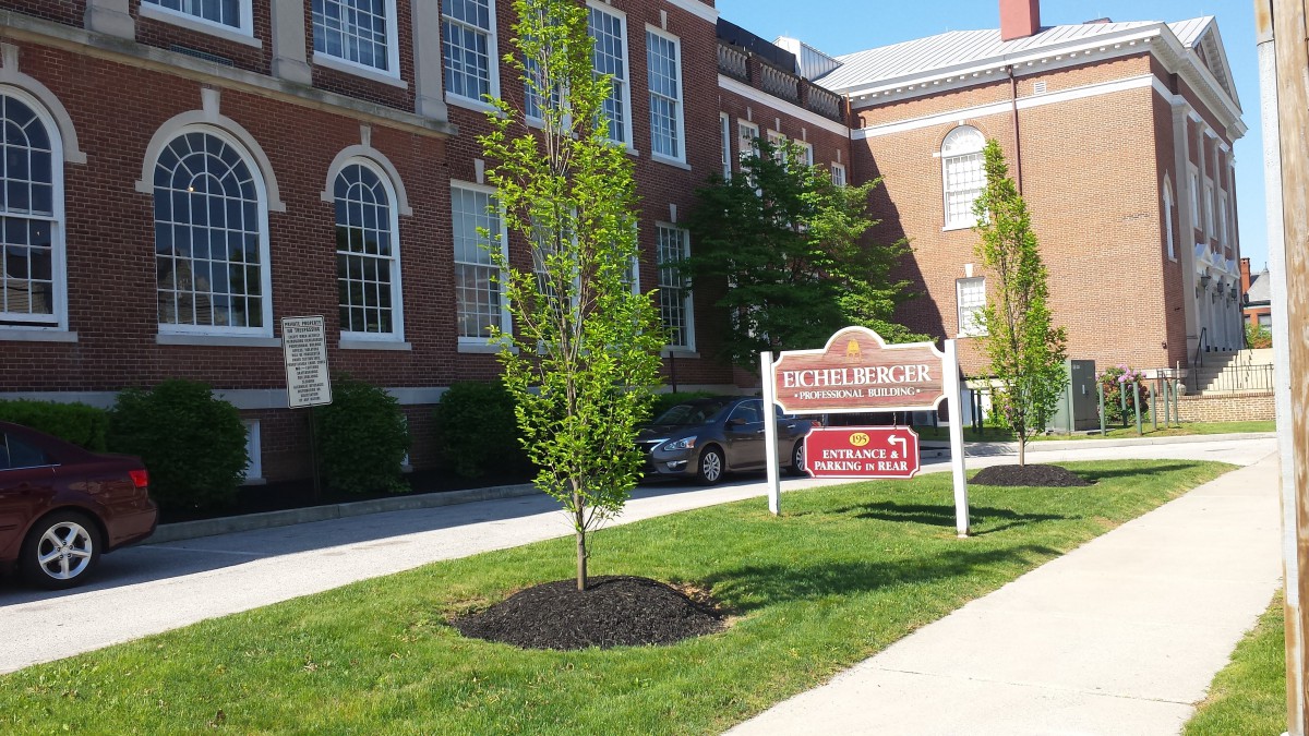 A brick building with a lawn, tree, and sidewalk next to a sign and parked car.