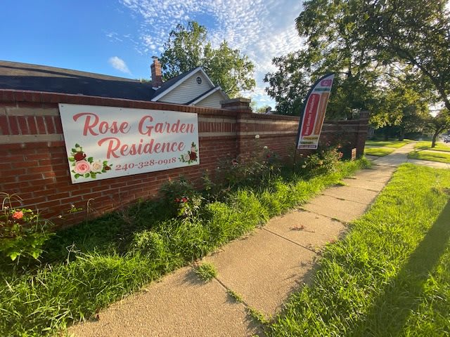 A residential area with a sign reading 'Rose Garden Residences' along a sidewalk on a sunny day.