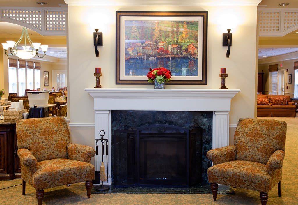 A cozy living room with a fireplace flanked by two armchairs, adorned with a painting and flowers.