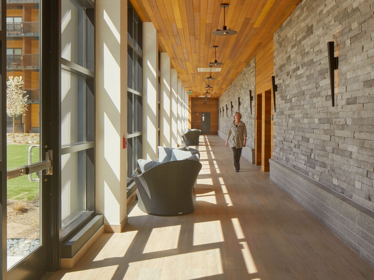 A modern hallway with large windows, stone walls, and wooden ceiling, featuring comfortable seating.