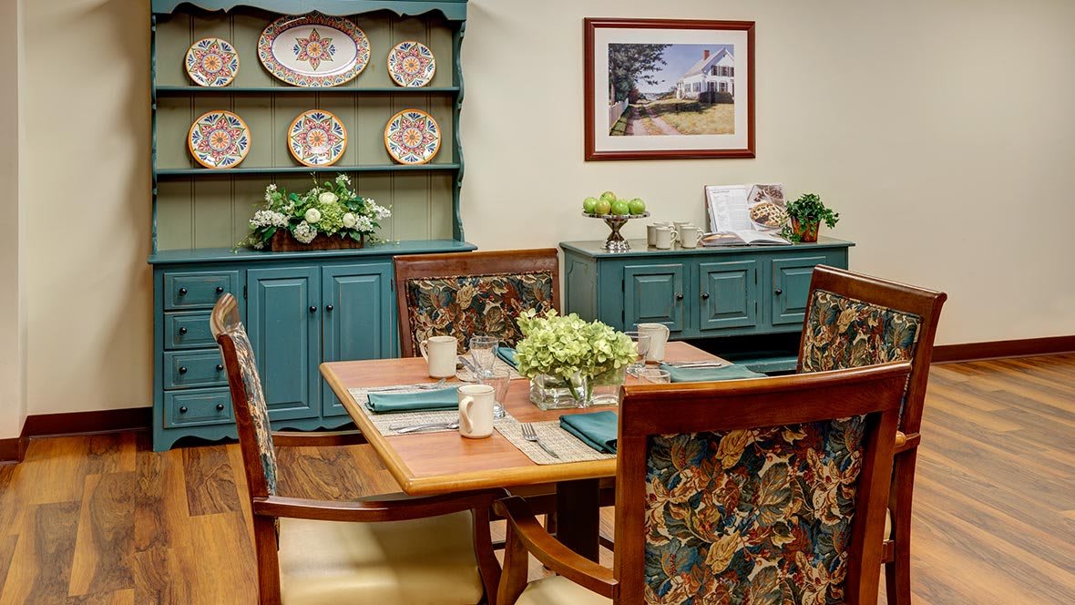 A cozy dining room with a wooden table, chairs adorned with floral patterns, and a china cabinet displaying decorative plates.