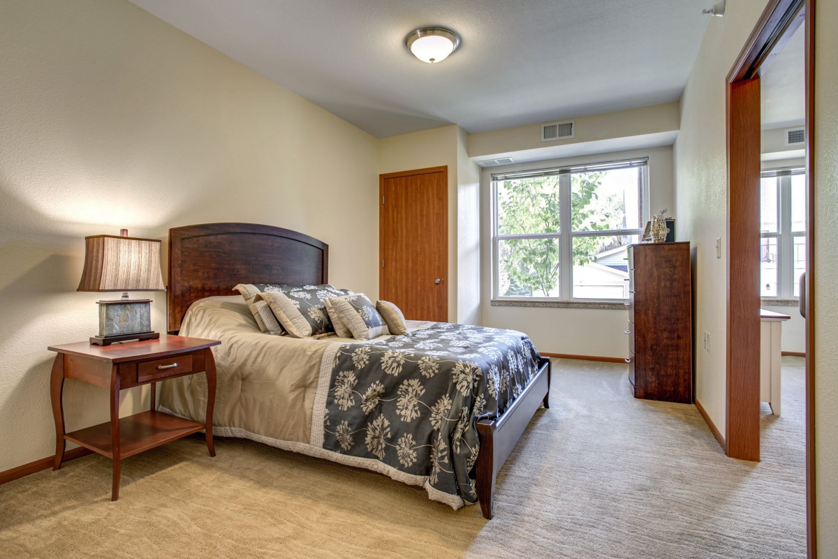 A cozy bedroom with a neatly made bed, wooden furniture, and a large window allowing natural light to pour in.
