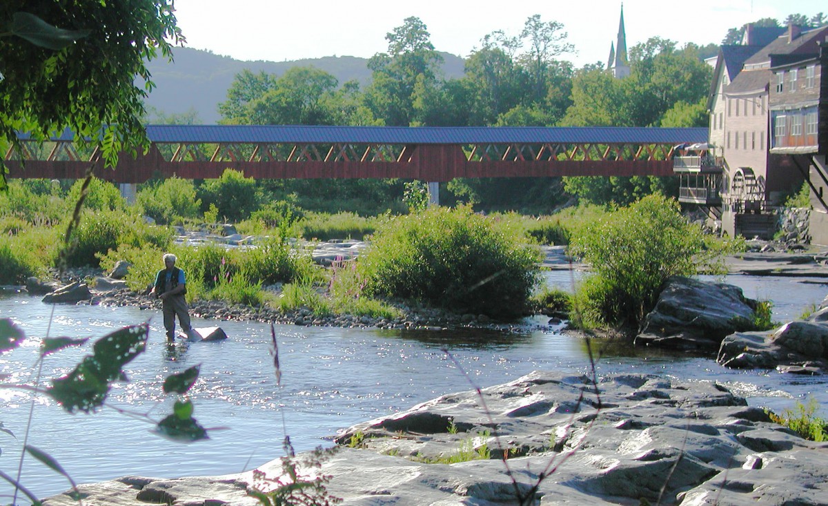A peaceful river scene with a person fishing near a picturesque covered bridge.