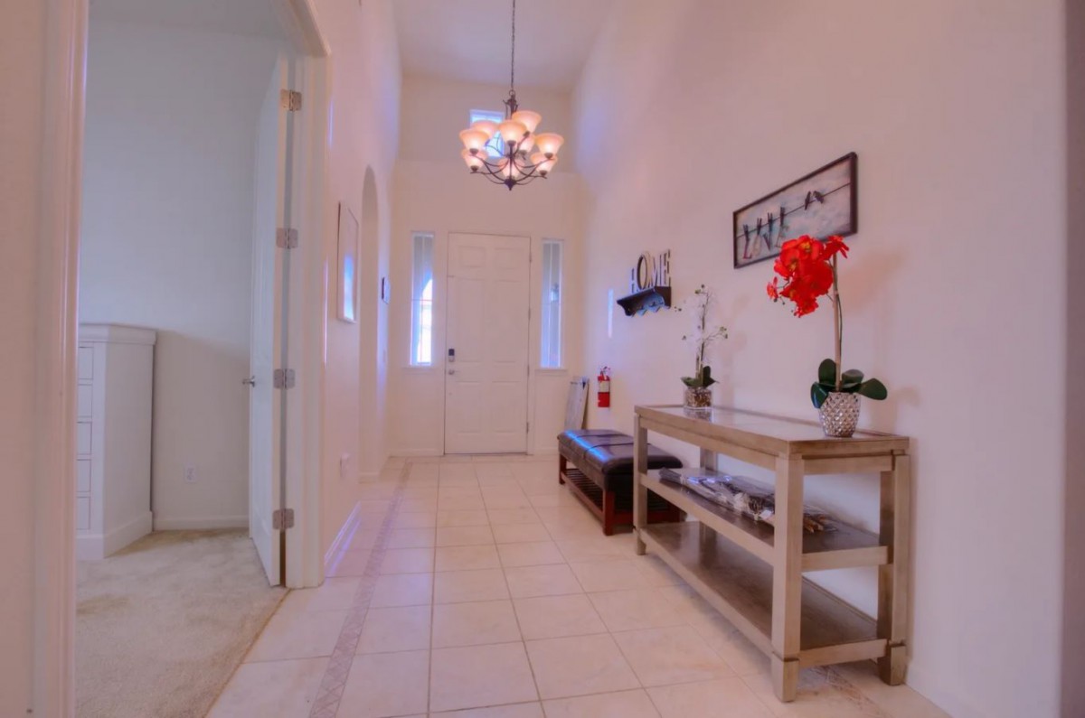 A bright hallway with white walls, a chandelier, and a wooden console table adorned with plants.
