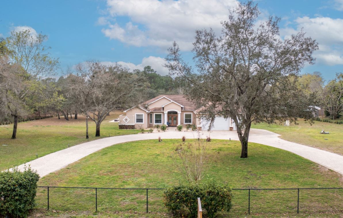 A suburban house with a circular driveway, surrounded by trees and a fenced yard under a partly cloudy sky.