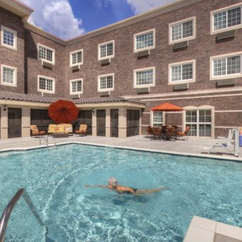 An outdoor swimming pool area at a hotel, featuring tables with umbrellas and clear blue skies above.