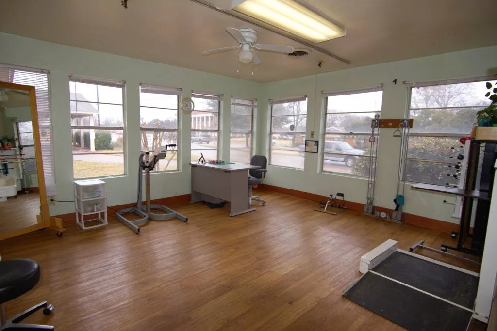 An empty physical therapy room with equipment, a desk, and large windows providing natural light.