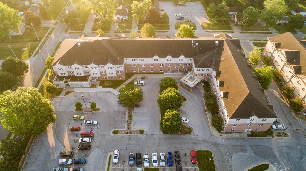 An aerial view of a building with a parking lot and surrounding trees under soft sunlight.