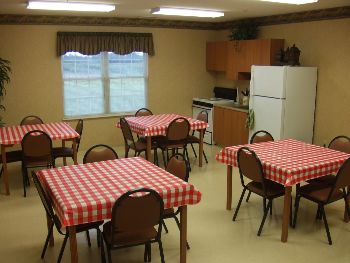 A dining area with multiple tables covered in red checkered tablecloths, surrounded by brown chairs. The room includes a small kitchen space with a refrigerator and microwave.