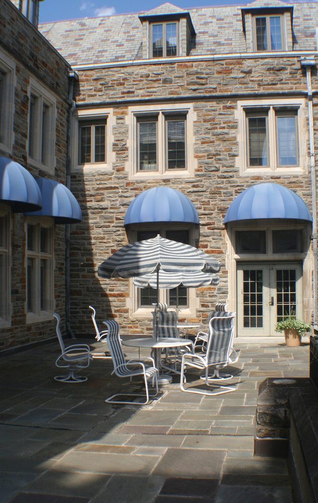 A quaint courtyard featuring outdoor seating with a striped umbrella, surrounded by a stone building with blue awnings.