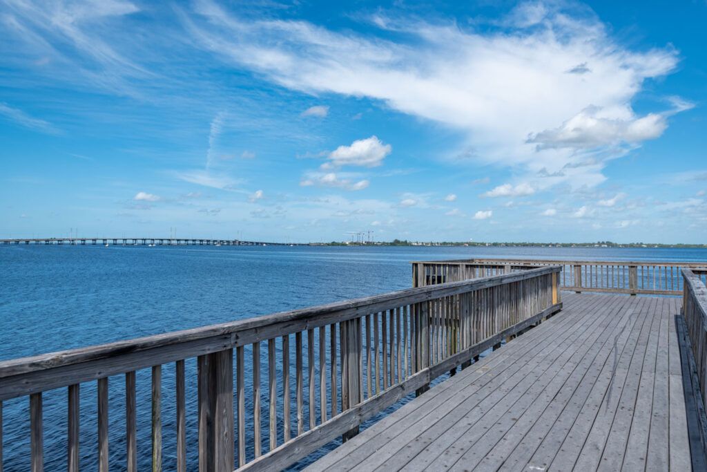 A serene view of a wooden pier extending over calm ocean waters under a blue sky with scattered clouds.