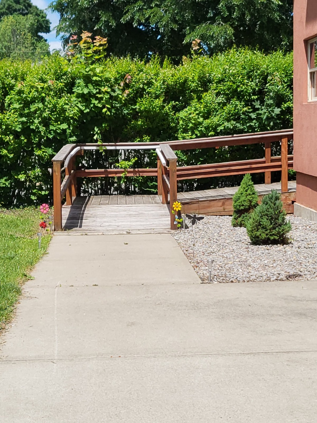A small wooden bridge connecting a sidewalk to a garden area, surrounded by greenery.