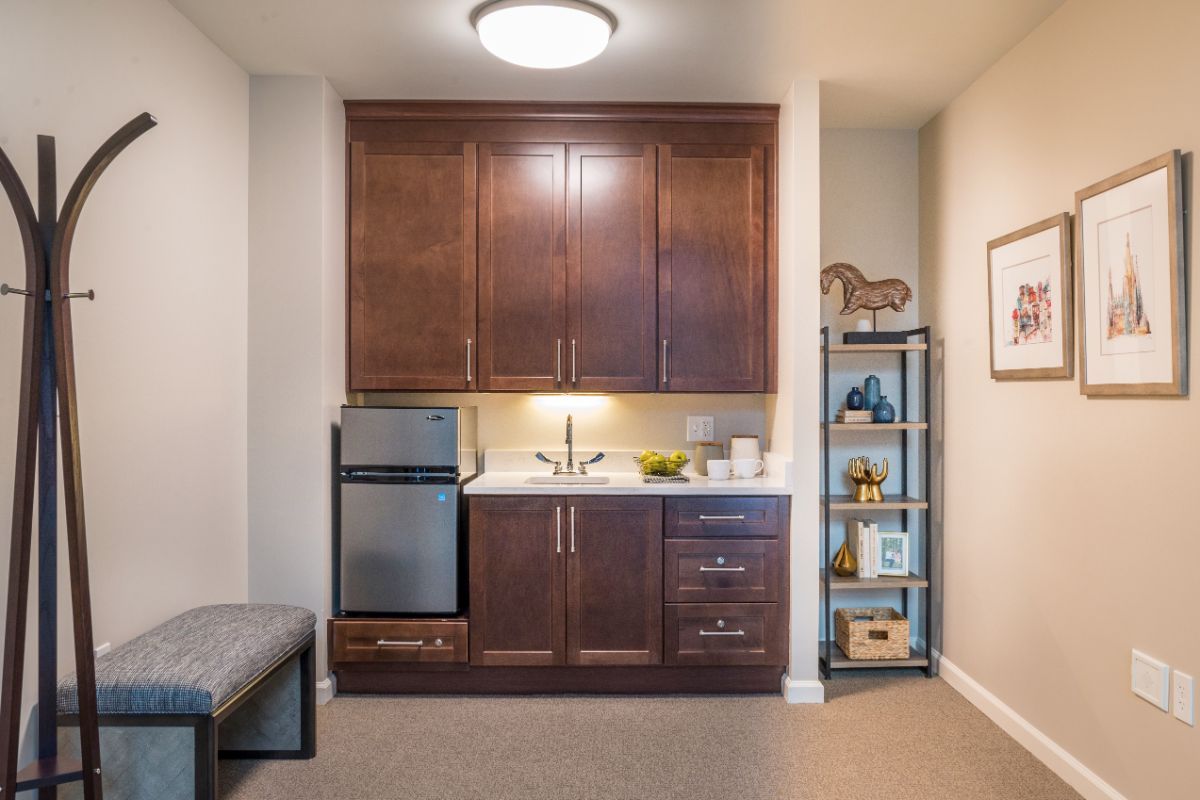 A compact kitchenette with dark wood cabinets, a mini refrigerator, and open shelving displaying decor items.