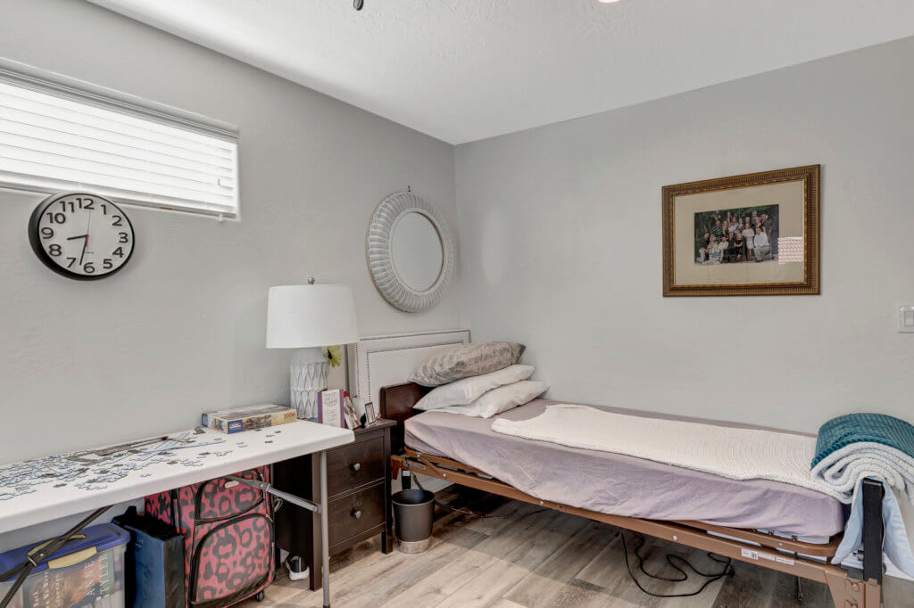 A minimalist bedroom with a single bed, featuring gray walls and wooden flooring, is simply decorated with a lamp, mirror, and framed picture.