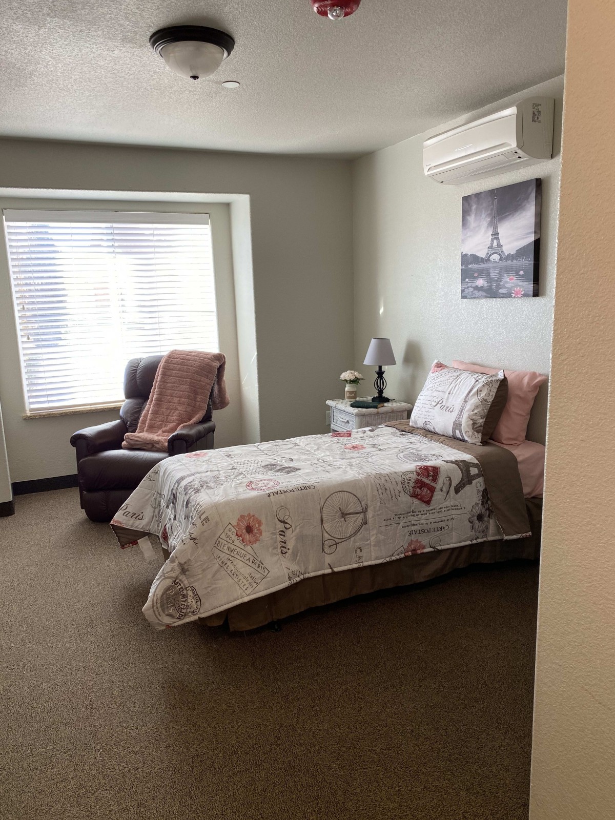 A cozy bedroom with a single bed, an armchair by the window, and a nightstand with a lamp.