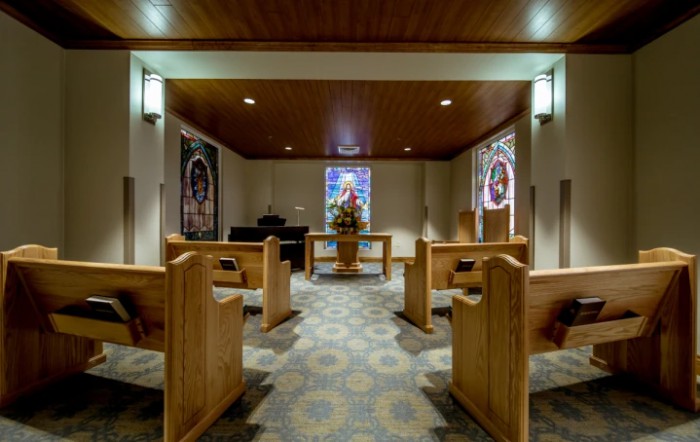 A serene chapel interior featuring wooden pews and a stained glass window at the altar.