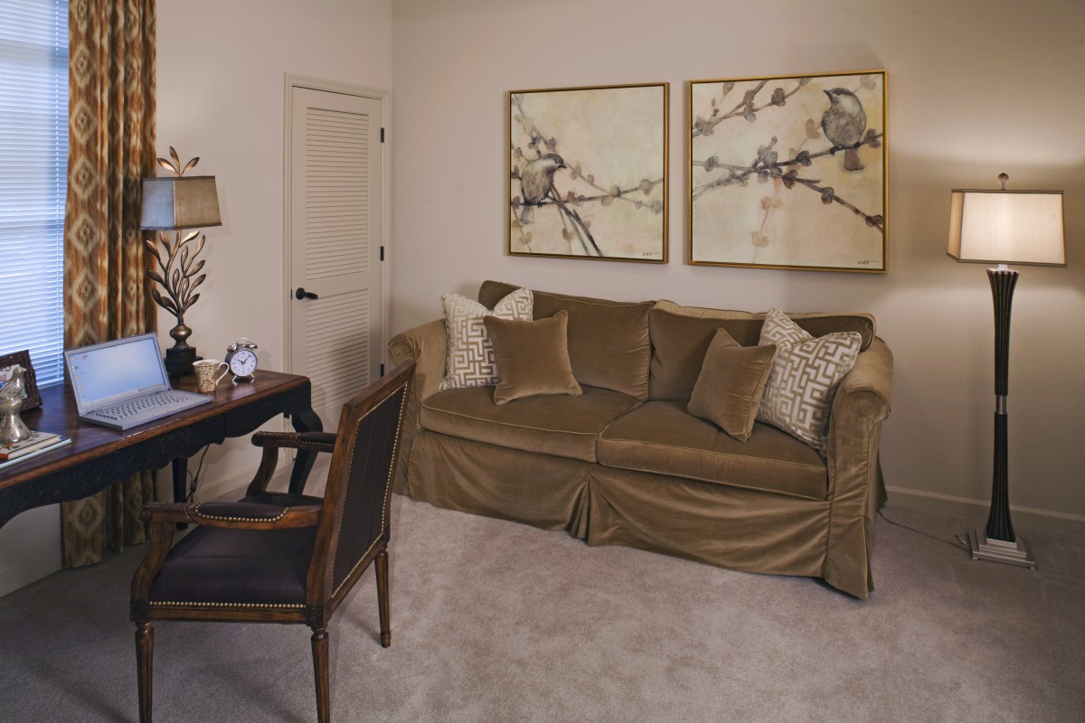A cozy living room with a brown sofa, decorative pillows, and abstract artwork on the wall. A desk with a lamp is positioned nearby.