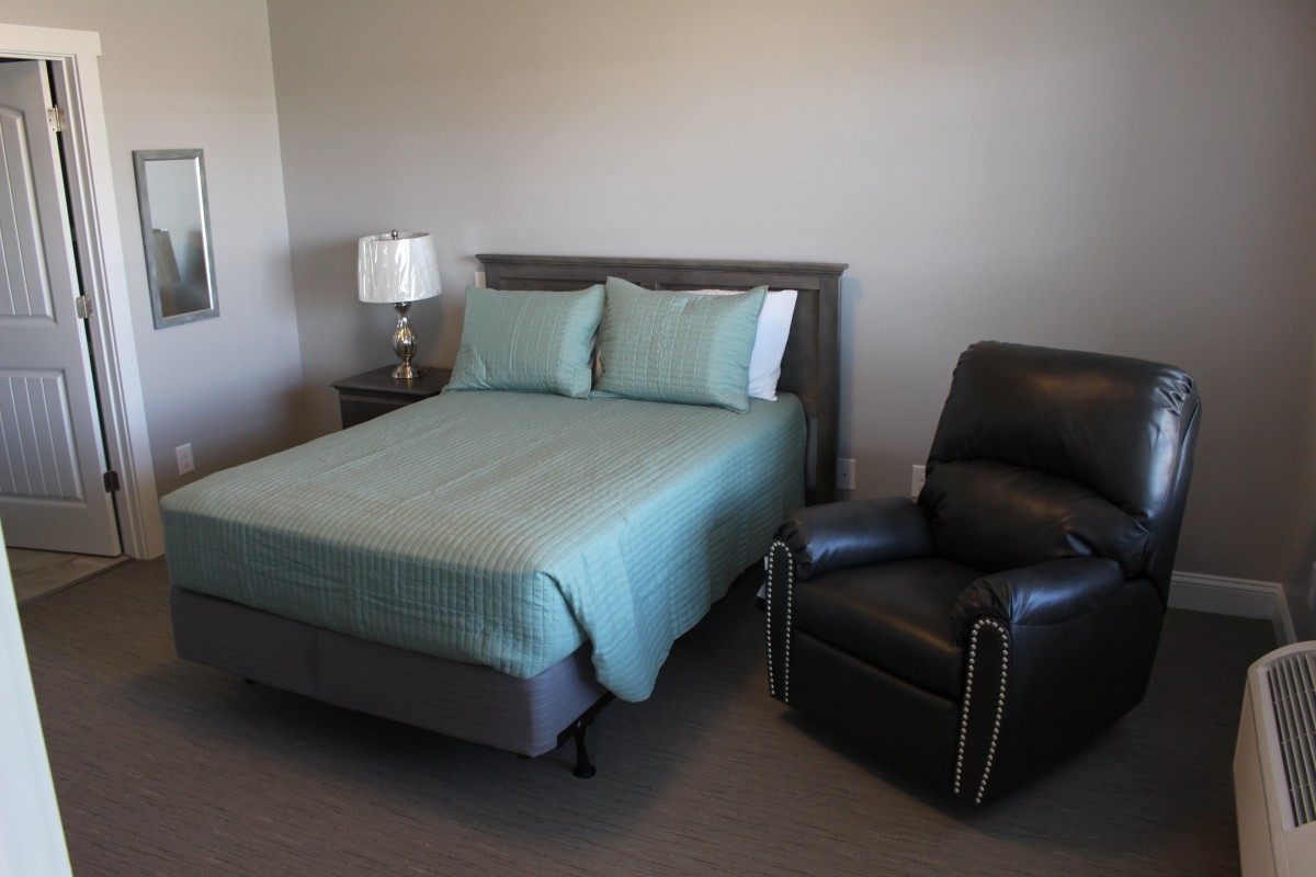 A cozy bedroom with a neatly made bed, a recliner chair, and a nightstand with a lamp.