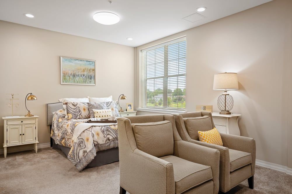 A cozy and well-decorated bedroom with neutral tones, featuring a bed, two armchairs, and a table lamp by the window.