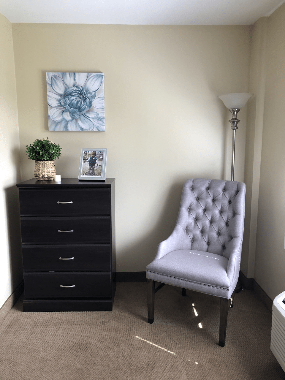 A cozy corner with a tufted chair, dresser, and floor lamp next to wall art.