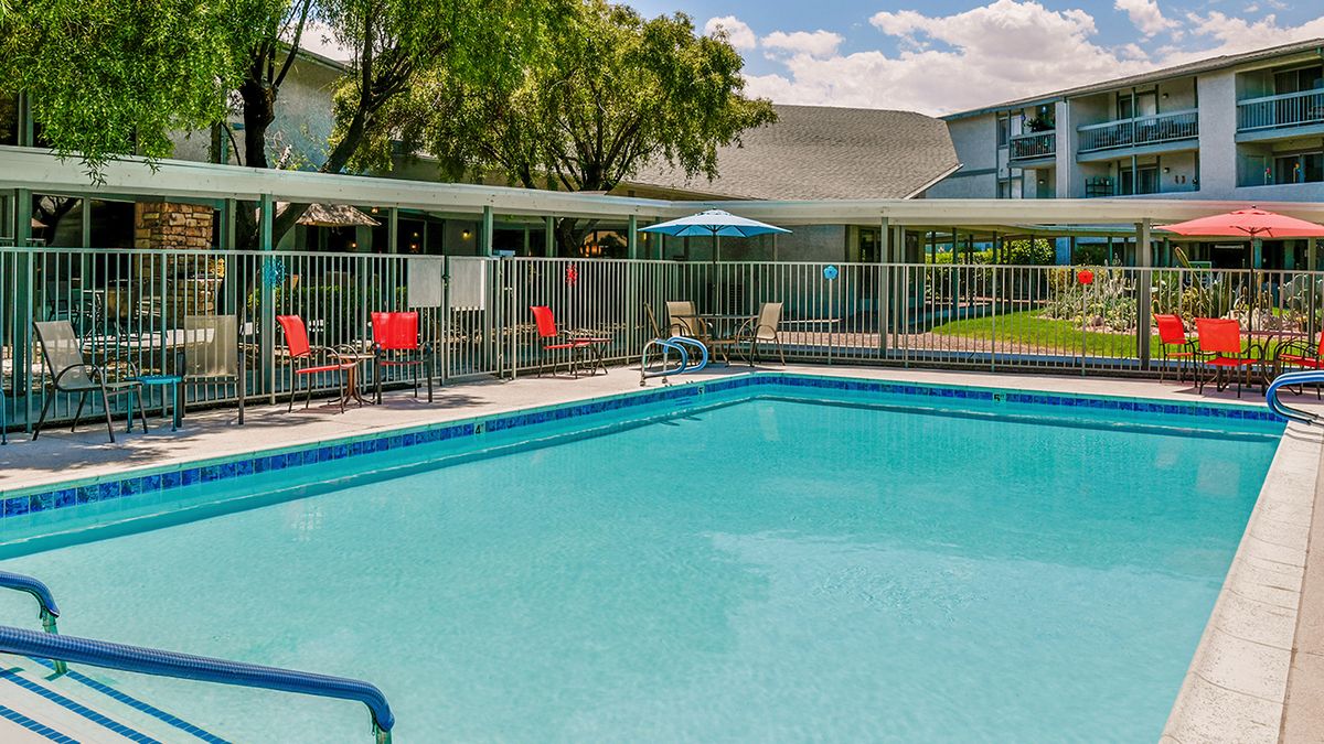 An outdoor swimming pool surrounded by a fence with chairs and an umbrella nearby.