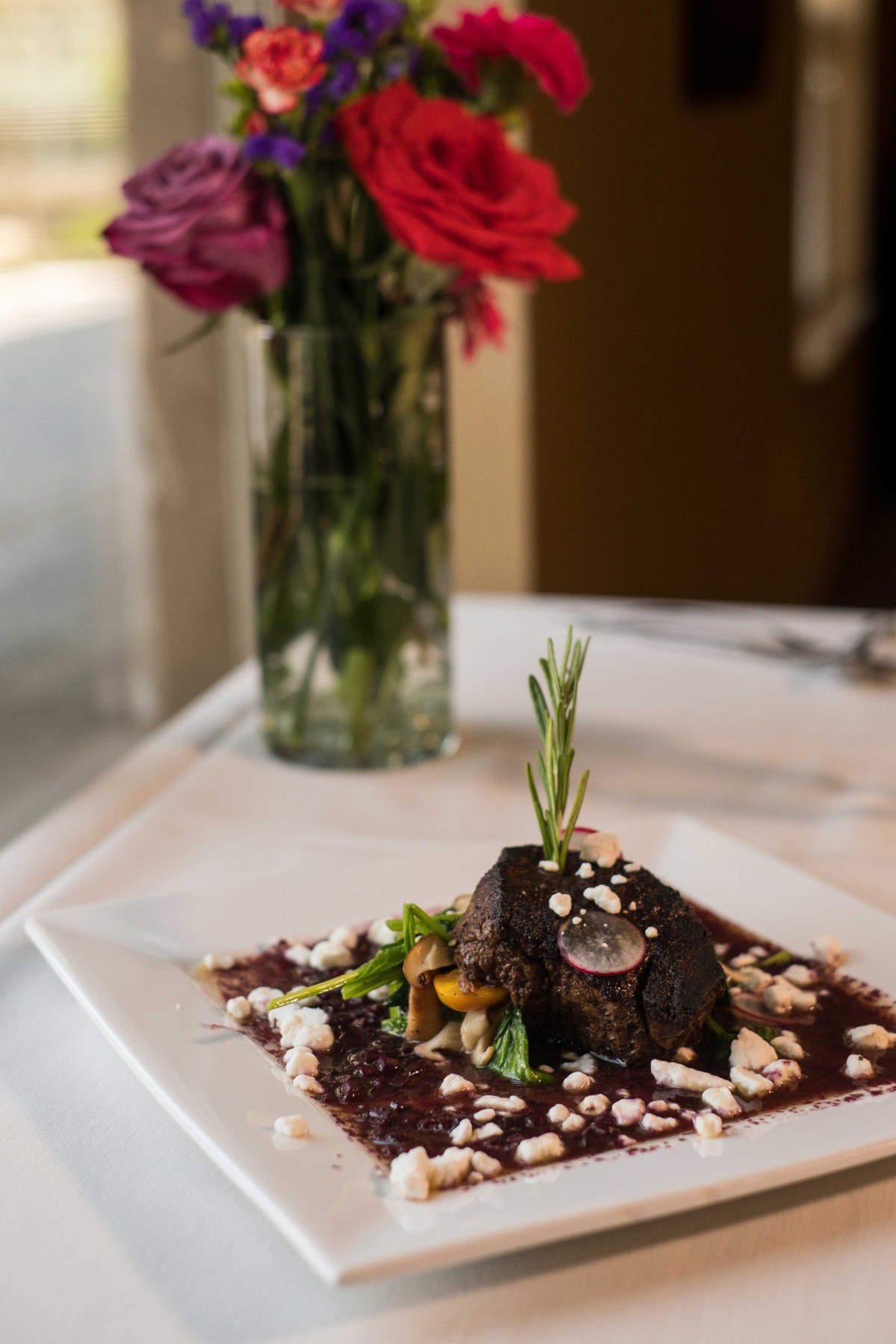 A beautifully plated gourmet dish served at an elegant restaurant, with a floral centerpiece in the background.