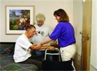 A caregiver assists an elderly man with physical therapy while another person observes.