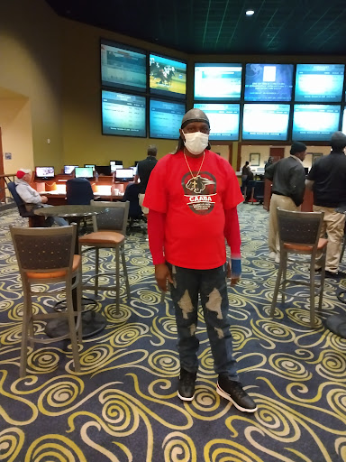 A person stands in a gaming area, wearing a red sweatshirt and a mask, with multiple sports screens in the background.