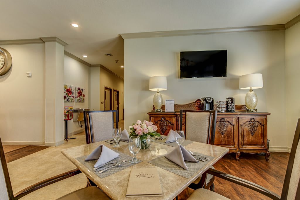 A well-decorated dining room with a table set for four, complete with napkins and floral centerpiece, complemented by elegant furnishings and a wall-mounted television.