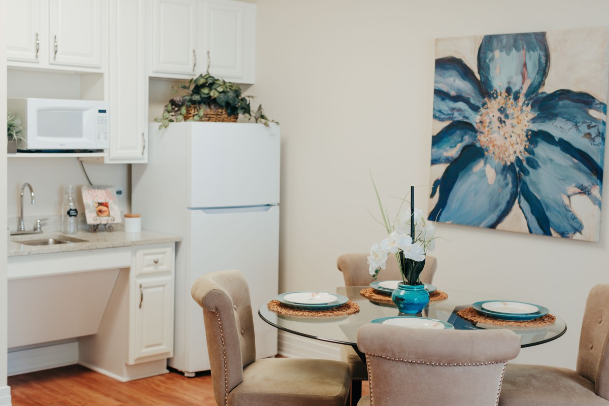 A cozy dining area with a round glass table, set with plates and a centerpiece, and a large floral artwork on the wall.