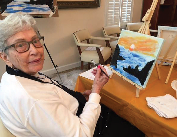 An elderly woman painting a scenic landscape on a canvas indoors.