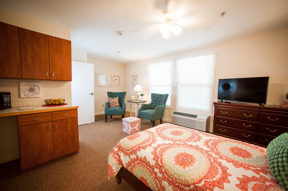 A cozy bedroom with a stylish bedspread, two comfortable chairs, a small round table, and a television near the window.