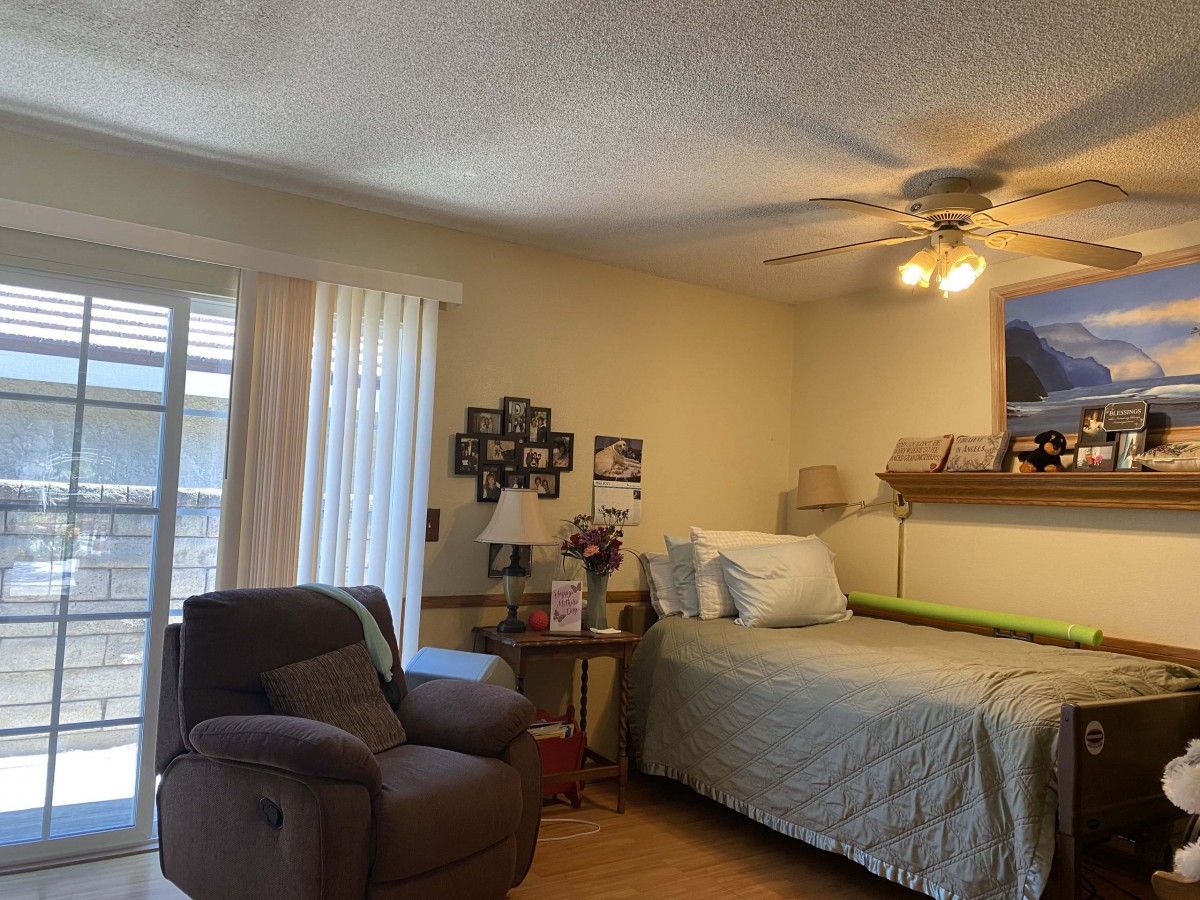 A cozy bedroom with a single bed, an armchair, and a ceiling fan, illuminated by natural light from a window.