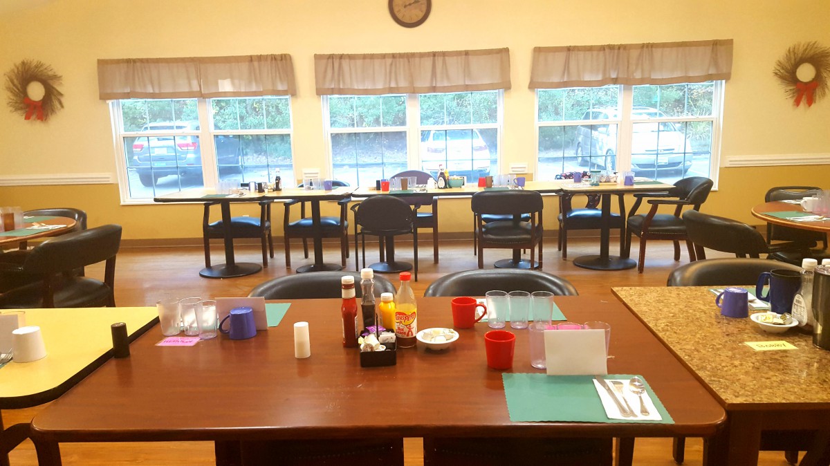 A dining area with neatly set tables and chairs, illuminated by natural light from large windows.