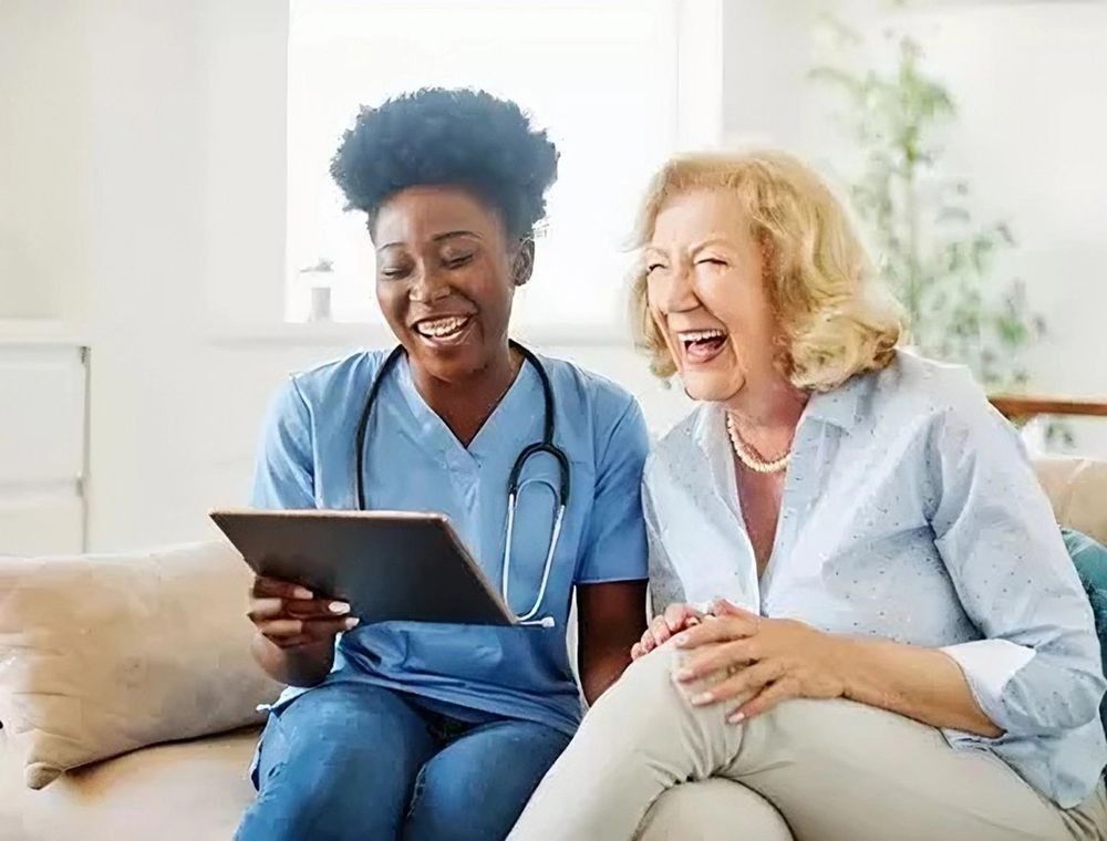 A nurse and an elderly woman share a joyful moment while looking at a tablet.