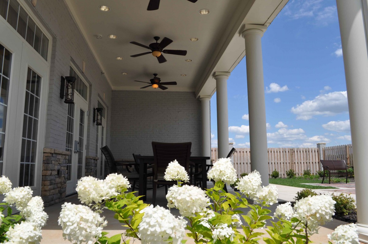 A cozy patio with overhead ceiling fans and a view of blooming white flowers against a cloudy sky.