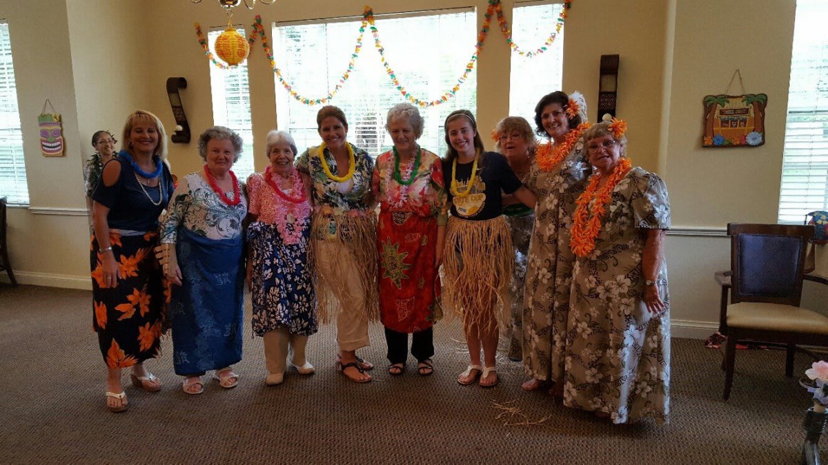 A group of women dressed in Hawaiian-themed costumes, wearing colorful lei garlands, posing together at an indoor party.
