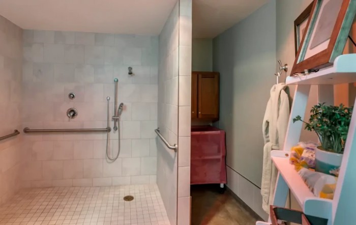 A bathroom with a walk-in shower featuring grab bars and tiled walls, alongside a bathrobe hanging near pastel-colored decor.