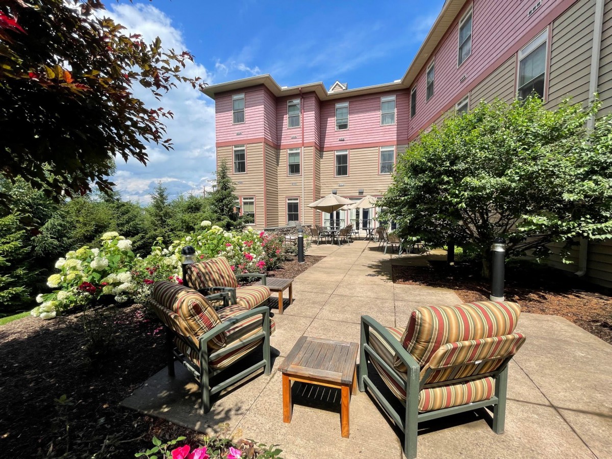 A comfortable outdoor patio area with seating, surrounded by lush greenery and set against a backdrop of a large building.