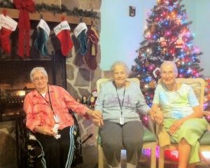 Three elderly people sitting in front of a decorated Christmas tree and fireplace, enjoying the holiday season together.