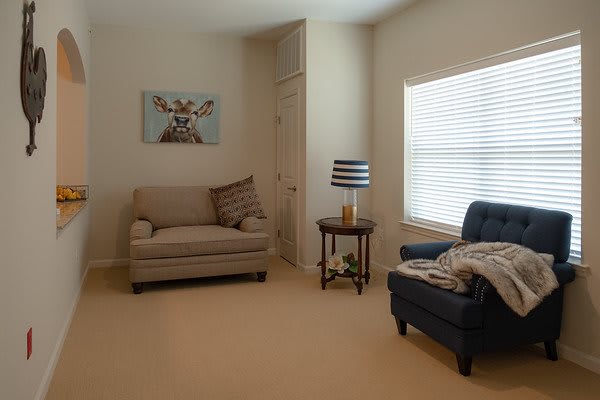 A cozy living room with a sofa, chair, and a side table featuring a lamp, complemented by wall artwork and natural light from the window.