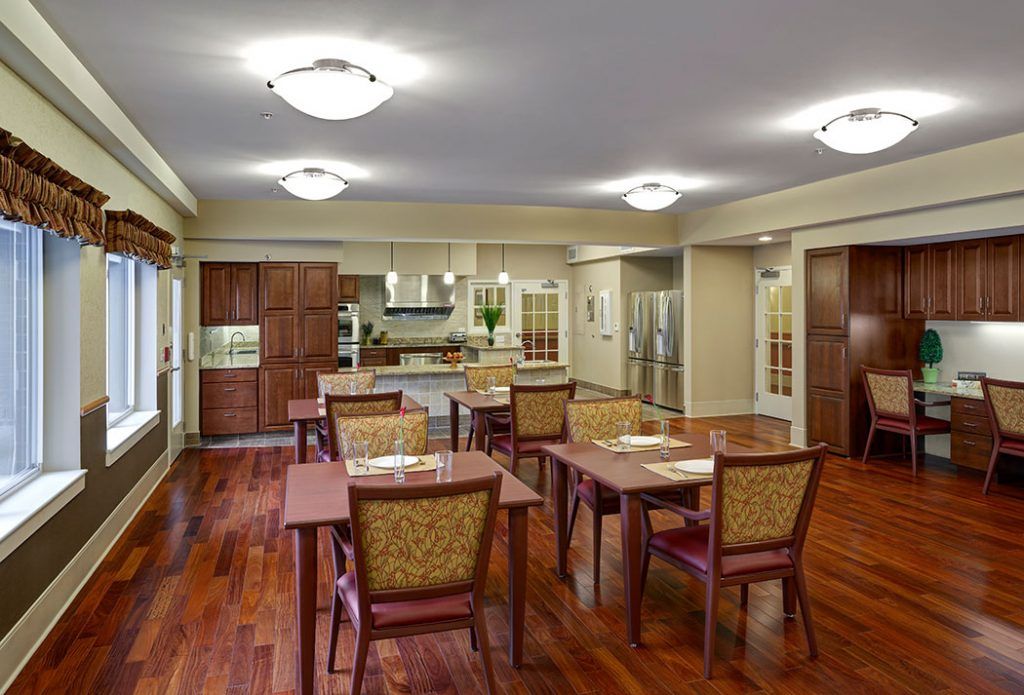 A spacious and modern dining area with wooden flooring and well-arranged tables and chairs, adjacent to a kitchen.
