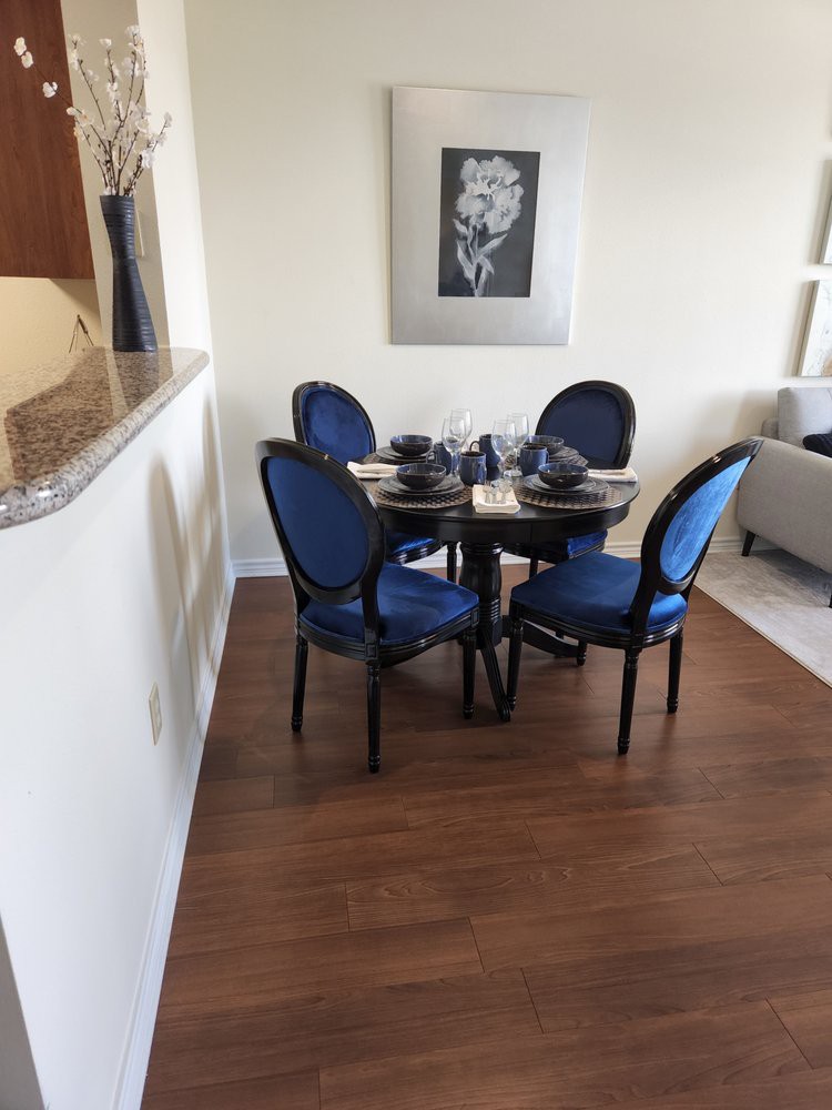 A cozy dining area features a round table set with elegant blue chairs and a simple table setting, complemented by artwork on the wall and hardwood flooring.